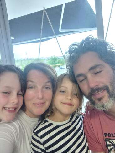 A happy family of four poses closely together, smiling warmly. The background shows a window and outdoor view. Their expressions reflect love, joy, and togetherness.