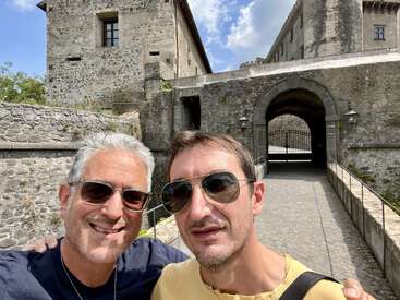 Two men pose for a selfie, smiling in front of a historic stone castle entrance with an arched gate, under a sunny sky with scattered clouds.