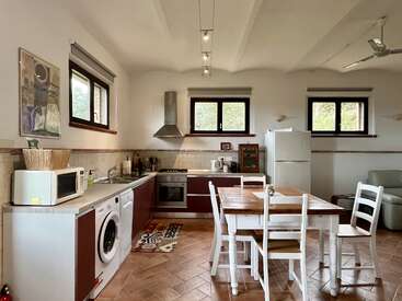 Diese gemütliche Küche verfügt über moderne Geräte, Holzschränke, einen Esstisch mit weißen Stühlen, Fliesenboden, große Fenster, einen Kühlschrank, eine Waschmaschine und eine zeitgemäße Einrichtung.