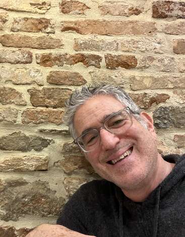 Un hombre sonriente, de pelo canoso y con gafas, posa delante de una pared rústica de ladrillo, vestido con una sudadera con capucha oscura. El ambiente es informal y alegre.