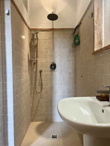 En esta imagen se ve un cuarto de baño moderno con azulejos beige, una ducha a ras de suelo con cabezal de ducha tipo lluvia, una ducha de mano, una repisa metálica y un lavabo de cerámica blanca.