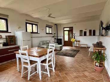 Esta habitación cuenta con una acogedora zona de estar con sofá, estufa de leña, mesa de comedor, cocina moderna, suelo de baldosas, varias ventanas, plantas y una decoración sencilla y rústica.