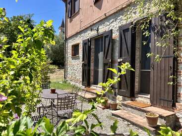 Una encantadora casa rústica de piedra con contraventanas de madera oscura, rodeada de vegetación. En el exterior hay una mesa y sillas de metal, perfectas para disfrutar de tranquilas tardes soleadas.