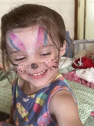 A young girl with bunny face paint smiles joyfully. She has pink and blue painted ears, a black nose, whiskers, and wears a colorful dress.