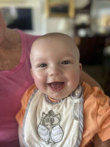 A smiling baby with bright eyes and a bib featuring an owl design sits on someone's lap, wearing an orange shirt, radiating happiness and joy indoors.