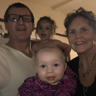 A smiling family of four poses together indoors: two adults, a young girl making a playful face, and a baby in front, all appearing happy and close.