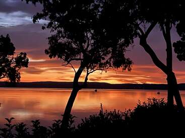 The image captures a serene sunset over a calm lake, with silhouettes of trees framing the vibrant orange, purple, and yellow sky reflected on the water.