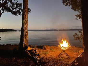 A glowing campfire burns near a tranquil lake at dusk, surrounded by trees and soft orange light, creating a serene and peaceful outdoor atmosphere.