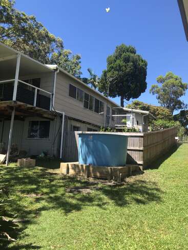 A sunny backyard with green grass, a blue water tank, wooden fencing, and a house with a covered porch. Tall trees and clear blue sky.