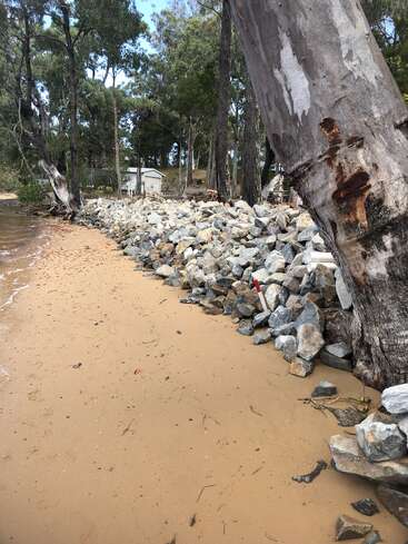 A sandy shoreline meets a rocky barrier, bordered by tall trees. In the background, a small white building sits among dense forest, creating a tranquil scene.