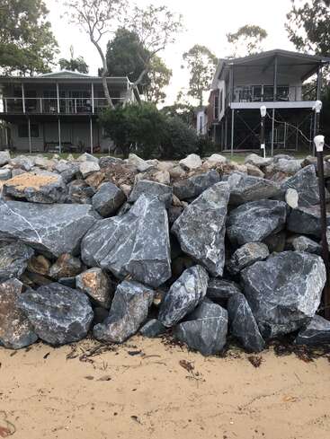 Large gray rocks form a protective barrier above sandy ground. Behind, two elevated houses stand surrounded by trees and greenery under a cloudy sky. Peaceful atmosphere.
