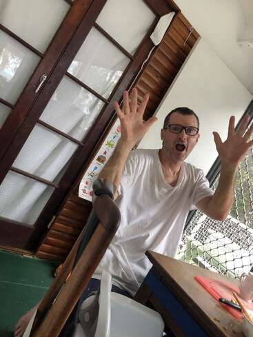 A man wearing glasses and a white shirt sits indoors, raising both hands with a surprised expression. There is a wooden door, mesh fence, and table nearby.