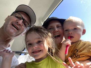 A cheerful family selfie with two adults and two young children, smiling brightly in sunlight. One child holds a red object, possibly a toy or straw.