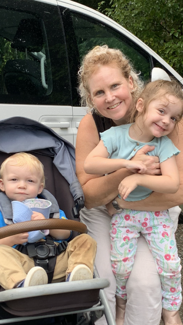 A smiling woman hugs a happy little girl while a blonde toddler sits in a stroller beside them, with a white car and greenery in the background.