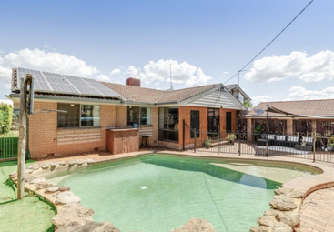 This image shows a cozy backyard with a swimming pool, brick house, solar panels, patio seating, safety fencing, clear blue sky, and some lush greenery.