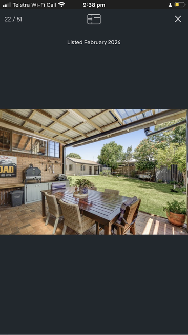 A covered patio with a wooden dining table and wicker chairs overlooks a spacious backyard, featuring green grass, a barbecue, potted plants, and outdoor kitchen.