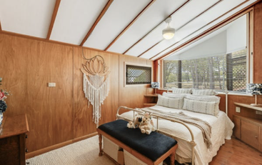 Cette chambre confortable est dotée de murs lambrissés, d'un lit blanc orné de coussins, d'un banc noir, de décorations murales en macramé, de grandes fenêtres et d'un ventilateur de plafond. Une ambiance chaleureuse et sereine.
