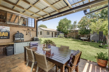Une terrasse couverte, équipée d'une table à manger en bois et de chaises en osier, donne sur un grand jardin où l'on trouve une pelouse verte, un barbecue, des plantes en pot et un coin cuisine extérieur.