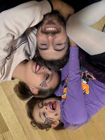 Un joyeux trio familial est allongé par terre, les visages tout près les uns des autres. Ils ont le visage peint en forme de toile d'araignée, arborent de larges sourires et portent des tenues sur le thème d'Halloween, partageant ainsi un moment de bonne humeur.