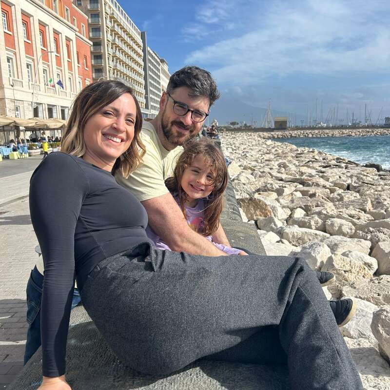 Une joyeuse famille de trois personnes est assise sur un mur de pierre au bord de la mer, souriant sous un ciel ensoleillé, avec les immeubles de la ville et le front de mer en arrière-plan.