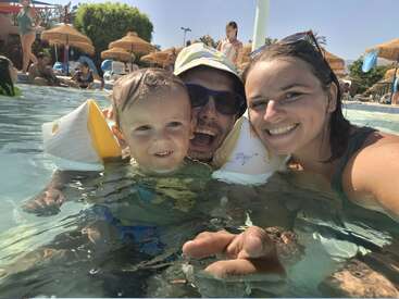 Uma família feliz desfruta de um dia ensolarado na piscina. Uma mulher, um homem e uma criança sorriem juntos na água, cercados por outros nadadores e guarda-chuvas.