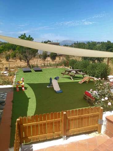 Um playground no quintal com grama artificial, escorregador para crianças, bancos, mesas de piquenique, brinquedos, guarda-sol, portão de madeira e vegetação exuberante sob um céu azul brilhante.