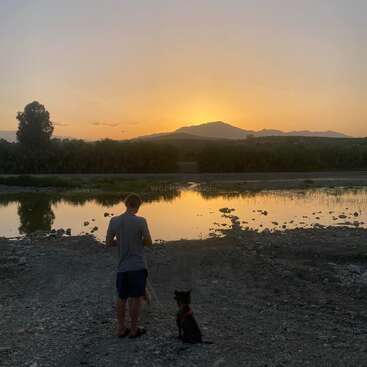 Une personne se tient à côté d'un chien près d'une rivière calme au coucher du soleil. Le ciel est orange, les montagnes et les arbres se reflètent dans l'eau paisible.