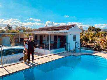 Un couple souriant se tient au bord d'une piscine sous un ciel bleu vif, avec une maison blanche et de la verdure en arrière-plan, profitant d'une journée ensoleillée.