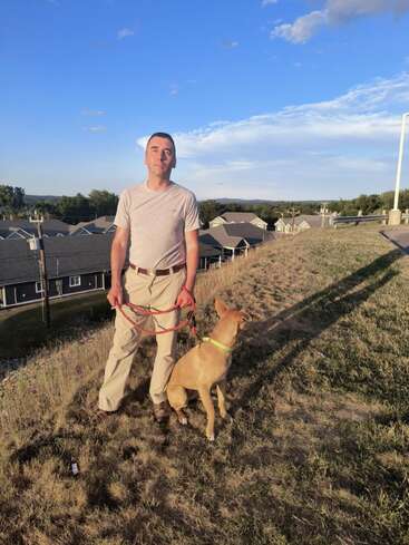 Un homme se tient sur une colline herbeuse en tenant la laisse d'un chien. Le chien est assis à côté de lui, des maisons et des arbres en arrière-plan, sous un ciel bleu.