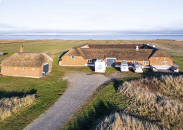 La imagen muestra una gran casa de dos plantas con tejado marrón y ribetes blancos, situada en un campo cubierto de hierba cerca del océano, rodeada de algunos vehículos.