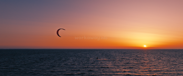 Un kitesurfeur glisse sur les vagues calmes de l'océan sous un ciel orange vif au coucher du soleil, créant une scène à la fois paisible et aventureuse à l'horizon.