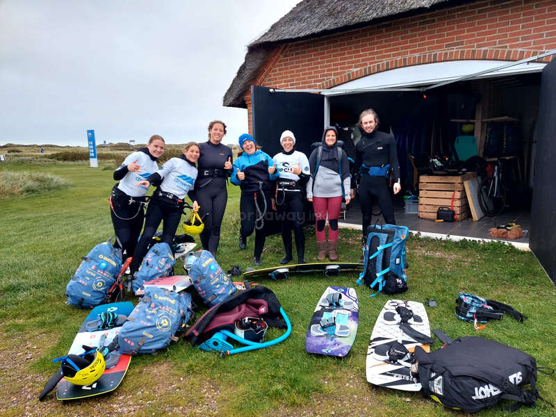 La imagen muestra a un grupo de personas de pie frente a un edificio de ladrillo, rodeadas de material de kitesurf, con un campo de hierba y un cielo nublado de fondo.
