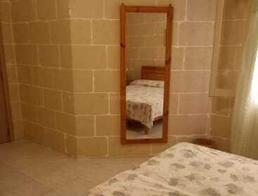 The image shows a simple bedroom with stone block walls, a wooden-framed mirror reflecting a bed, and a window with white curtains letting in natural light.