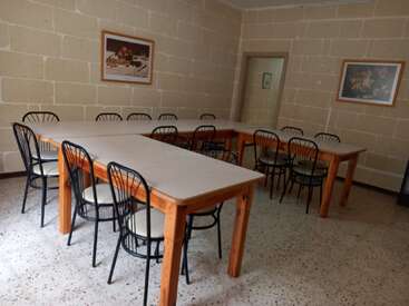 This image shows a simple, empty dining room with beige stone walls, several rectangular tables arranged in a U-shape, black chairs, and framed wall art.