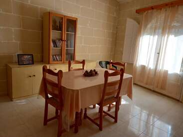 A sunlit dining room with a wooden table, four chairs, glass-front cabinet, sideboard, sheer curtains, tiled floor, and neutral stone walls. Simple, cozy, inviting atmosphere.