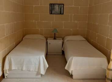 The image depicts a small, simple bedroom with two single beds, a nightstand with a lamp between them, bare stone walls, and a framed picture above.