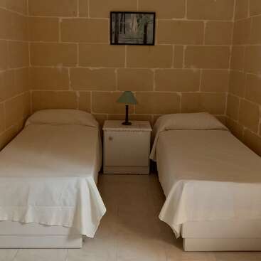 L'image représente une petite chambre simple avec deux lits simples, une table de nuit avec une lampe entre les deux, des murs en pierre nue et un tableau encadré au-dessus.