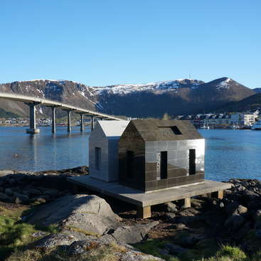 La imagen representa una escena serena de una pequeña estructura en forma de casa en una costa rocosa, con un puente sobre una masa de agua y montañas al fondo.