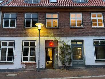 The image depicts a two-story building with a red brick facade, white walls, and a tiled roof, featuring windows and a doorway with a red light fixture.