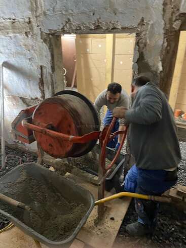 Two men work in a construction site, one operating a cement mixer, the other standing beside it, amidst a backdrop of concrete walls and a wheelbarrow filled with cement.