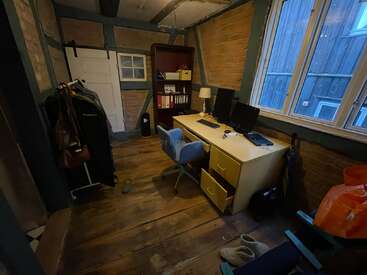 The image depicts a cluttered room with a desk, chair, and window, featuring wooden floors and walls, with various items scattered about, including a coat rack and bookshelf.