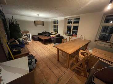 The image depicts a cluttered room with wooden floors and furniture, featuring a couch, table, chairs, and windows, with a dimly lit atmosphere suggesting a storage or living space.
