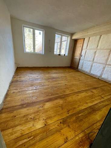 The image depicts an empty room with white walls, a wooden door, and a window, featuring a light-colored wood floor and a textured ceiling.
