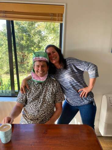 A man wearing a playful dinosaur hat sits next to a smiling woman indoors, both looking happy. A cup of coffee sits on the wooden table.