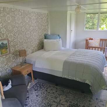 A cozy bedroom with a single bed, patterned wallpaper, nightstand, small lamp, window, armchair, framed picture, crib, soft rug, and natural light.