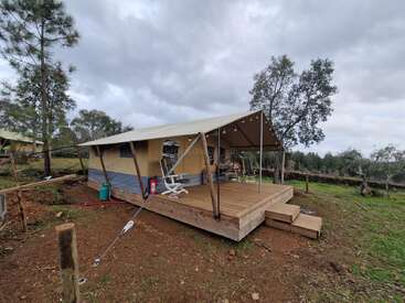 Une tente glamping confortable est installée sur une plate-forme en bois, entourée d'arbres et d'une pelouse sous un ciel nuageux, offrant un refuge confortable en plein air.