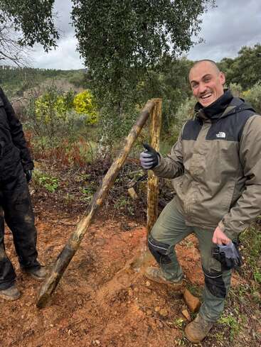 A smiling man in outdoor work clothes gives a thumbs up beside wooden fence posts. The ground is muddy, and trees fill the background. It looks like teamwork.