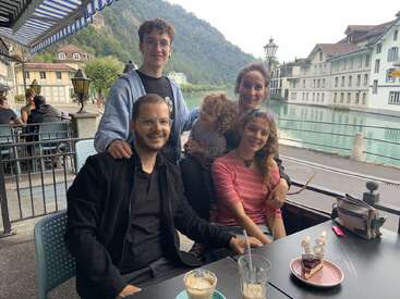 Eine fünfköpfige Familie sitzt in einem Straßencafé an einem ruhigen Fluss, umgeben von Bergen und charmanten Gebäuden. Sie lächeln und scheinen einen Geburtstag zu feiern.