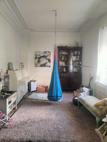 Ein gemütliches Wohnzimmer mit einem blauen Hängesessel, einem großen Teppich, einem Bücherregal, einem Klavier, Kinderspielzeug, einem alten Sofa, Kunstwerken und viel natürlichem Licht.