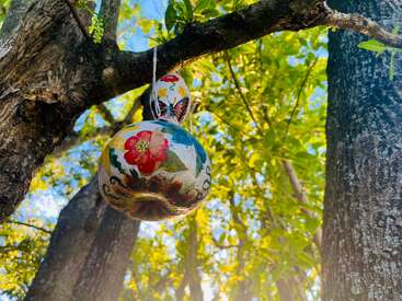 Ein handbemalter Kürbis mit bunten Blumen und einem Schmetterling hängt an einem Ast, umgeben von leuchtend grünen Blättern und dem Sonnenlicht, das durch die Baumkronen fällt.
